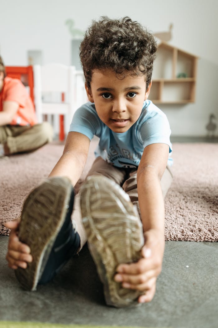 A young boy stretches his feet forward while sitting indoors in a kindergarten setting.